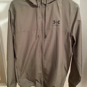 Under Armour träningsset i mörk grönt - Snyggt träningsset från Under Armour i mörk grönt, består av en lätt jacka med huva, dragkedja och snörning samt matchande byxor med resår i midjan. Perfekt för dig som gillar sportig och stilren look.