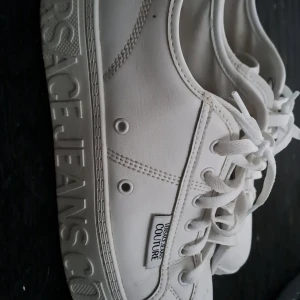 Vita sneakers från Versace Jeans Couture - Pris kan diskuteras.  Snygga vita sneakers från Versace Jeans Couture med tydlig logga på sidan och längs sulan. Skorna har klassisk snörning. Säljer mina nästan oanvända versache skor för de inte passar. 