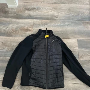 Svart hybridjacka från Parajumpers - Svart hybridjacka från Parajumpers med quiltad front och rygg i blankt material samt ärmar i stretchigt tyg. Jackan har hel dragkedja, bröstficka med dragkedja och hög krage. Perfekt för dig som gillar sportig och clean stil.