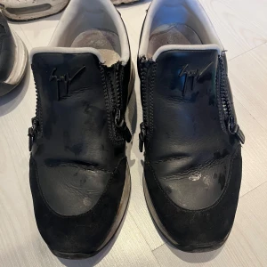 Svarta sneakers från Giuseppe Zanotti - Svarta sneakers från Giuseppe Zanotti med coola dubbla dragkedjor på ovansidan och ikonisk logga. Skorna har en mix av skinn och mocka, vit innersula och svart/vit sula. Perfekta för dig som gillar en edgy och stilren look.
