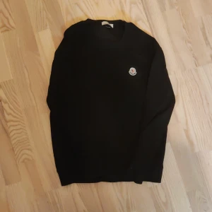 Svart Moncler tröja - Svart stickad tröja från Moncler med klassisk rund hals och broderad logga på bröstet. Långärmad modell med ribbade muddar och enkel, clean design. Perfekt för dig som gillar stilrena plagg med exklusiv känsla.