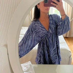 Blå mönstrad kortklänning med öppen rygg - Supersöt blå kortklänning med vitt mönster och v-ringning. Klänningen har långa, luftiga ärmar och en snygg öppen rygg med knytband. Aldrig använd!! Storlek xs men passar en xs-s!! Öppen för prisförslag