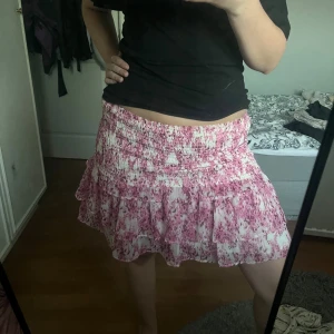 Rosa blommig volangkjol - Supersöt kort kjol med rosa och vita blommor, smockad midja och volanger. Lätt och luftig, perfekt för varma dagar. Kjolen har en feminin vibe och passar dig som gillar romantisk stil. Perfekt till både topp och kjol! 