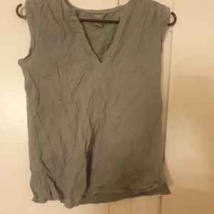 Grå ärmlös linnetopp från Lindex XS - Snygg grå ärmlös linnetopp från Lindex i storlek XS. Toppen har en avslappnad, lite skrynklig look och v-ringning framtill. Perfekt för en chill och stilren outfit, lätt att matcha med jeans eller kjol.