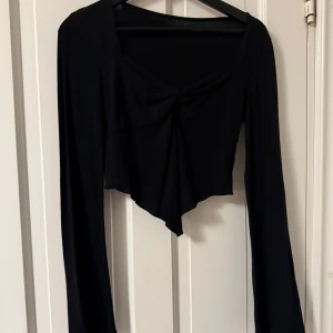 Svart croppad långärmad topp - Snygg svart croppad topp med lång ärm och twistad detalj framtill. Toppen har en spetsig nederkant och fyrkantig ringning, vilket ger en cool och trendig look. Perfekt att styla med höga jeans eller kjol.