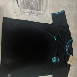 Svart Nike Brasilien fotbollströja  - Helt ny Svart fotbollströja från Nike med blå detaljer och CBF-emblem på bröstet. Tröjan är tillverkad i Dri-FIT-material som andas och har korta ärmar. Perfekt för dig som gillar stilren design och vill representera Brasilien på planen.