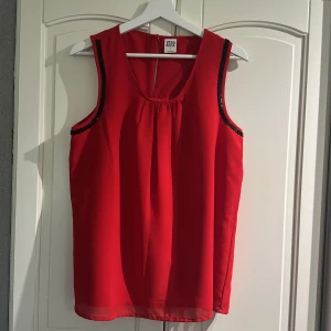 Röd ärmlös blus från Vero Moda - Snygg röd blus från Vero Moda i ärmlös modell med svarta paljett-detaljer längs ärmhålen. Blusen har rund halsringning med lätt rynk framtill och är tillverkad i ett tunt, flowy material som ger en luftig känsla. Använd ett fåtal ggr