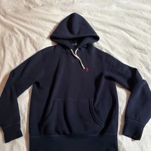 Mörkblå hoodie från Polo Ralph Lauren - Snygg mörkblå hoodie från Polo Ralph Lauren med klassisk huva och vit snörning. Tröjan har en röd broderad logga på bröstet och en stor magficka. Perfekt för dig som gillar stilrena och bekväma plagg med premiumkänsla. Mitt pris 599 nypris 2195