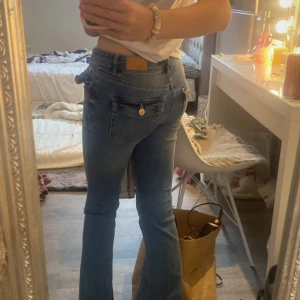 Blå bootcut jeans med fickdetaljer - Säljer ett par snygga blå bootcut jeans med coola fickor bak som har guldiga knappar. Jeansen har låg midja och klassisk femficksdesign. Materialet är jeans i bomull och de har en lätt utsvängd passform nertill. Perfekta för dig som gillar retrovibbar!