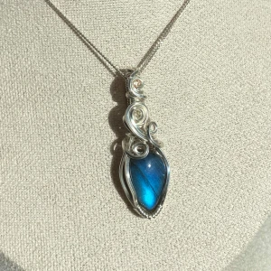 Silverberlock med labradorit  - Unik berlock till halsband med en glansig blå labradorit infattad i konstnärligt virad silverfärgad tråd. Hänget har en droppformad design med snygga snirkliga detaljer som ger en magisk och cool vibe. Perfekt för dig som vill sticka ut med något speciellt. Kedja ingår inte. Går att köpa till för 50kr.