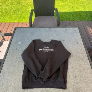 Svart sweatshirt från Peak Performance - Svart sweatshirt från Peak Performance med vit broderad logga på bröstet. Klassisk rund halsringning, ribbade muddar och nederkant. Perfekt basic-tröja för dig som gillar stilren och sportig look.