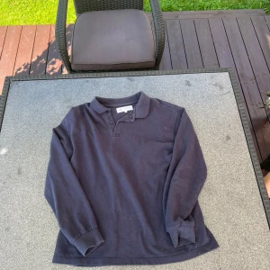 Mörkblå pikétröja från Zara - Klassisk mörkblå långärmad pikétröja från Zara med krage och knappar framtill. Tillverkad i mjuk bomull som känns skön mot huden. Perfekt för dig som gillar en enkel och stilren look med lite sportig känsla.