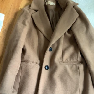 Klassisk beige kappa med knappar - Snygg beige kappa med två svarta knappar framtill och klassisk krage. Jackan har raka fickor och en stilren, tidlös design som passar perfekt till höst och vinter. Materialet känns mjukt och värmande, och modellen är normal i passformen.