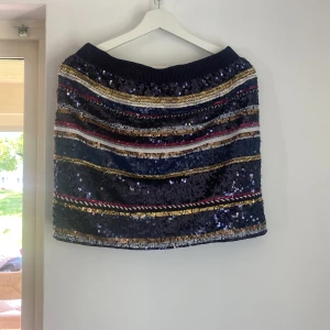 Paljettkjol från Holly & Whyte - Paljettkjol från Holly & Whyte med färgglada ränder i svart, guld, blått och rött. Kjolen har elastisk midja och är täckt av glittriga paljetter!