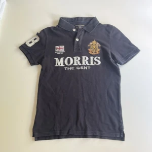 Svart pikétröja från Morris - Klassisk svart pikétröja från Morris Gents i 100% bomull. Tröjan har broderade detaljer, tryck på bröstet och ärm, samt krage med knappar. Snygg passform Storlek xs perfekt för en kille runt 14 år.