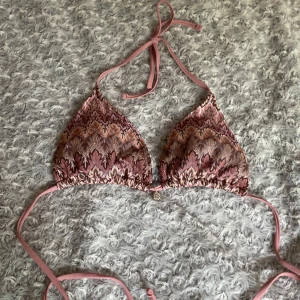 Rosa mönstrad bikini med snäckberlock - Supersöt trekantsbikini i rosa och bruna toner med broderat mönster. Bikinin har knyt i nacken och ryggen samt en liten snäckformad berlock framtill. Tillverkad i 100% polyester. Aldrig använd, helt ny. Storlek s.