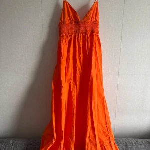 Orange långklänning från Zara - Superfin orange långklänning från Zara med smala axelband och smockad midja. Klänningen har v-ringning och är tillverkad i ett lätt bomullstyg, perfekt för varma dagar. Snyggt flowy fall och riktigt härlig färg som sticker ut.