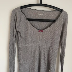 Grå ribbad tröja Odd Molly med rosett - Grå ribbad långärmad tröja från Odd Molly med v-ringning, svart spetskant och liten vinröd rosett framtill. Unik kompass- och hjärtdetalj på ärmen. Mjuk och stretchig bomullskvalitet, perfekt för lager-på-lager och chill vibes. Köpte för 350 aldrig använd.