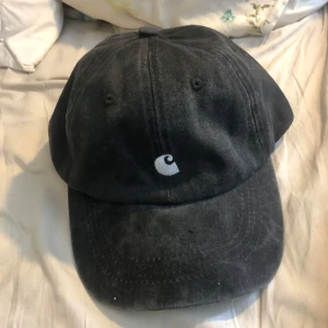 Svart Carhartt keps i bomull - Svart keps från Carhartt med klassisk böjd skärm och broderad logga framtill. Tillverkad i bomullstyg som ger en avslappnad och cool look.