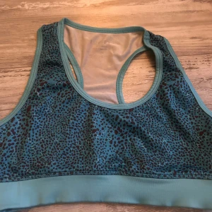 Turkos sporttopp med leopardmönster - Sportig turkos linnetopp från H&M med svart leopardmönster. Modellen är ärmlös med bred resår nedtill och brottarrygg. Perfekt för träning och ger en cool vibe med sitt djurmönster. Materialet är stretchigt och andas, vilket gör den bekväm under intensiva pass.