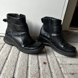 Svarta boots i skinn med dragkedja - Svarta boots i skinn med rund tå och platt sula. Snyggt stilrena med dragkedja på sidan och bred rem över vristen. Perfekta för höst och vinter när du vill ha både komfort och stil.