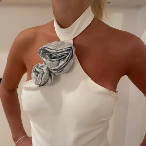 Vit halterneck topp med silverblommor - Unik vit halterneck topp med en axel och stora dekorativa silverblommor framtill. Toppen har hög hals med spänne i nacken och är axelbandslös på ena sidan. Perfekt för dig som vill sticka ut med en cool och elegant look.