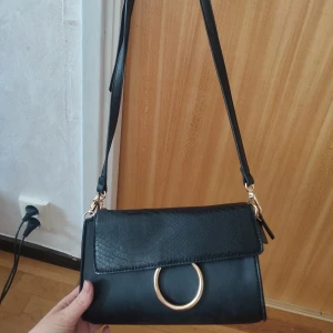 Svart axelväska med guldring - Snygg svart axelväska med justerbart axelband och lock med struktur. Väskan har en stor guldfärgad ring framtill som detalj och stängs med magnetknapp. Perfekt storlek för mobil, plånbok och nycklar.