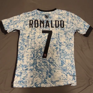 Portugal Ronaldo #7 Nike fotbollströja - Snygg Portugal fotbollströja från Nike med Ronaldo och nummer 7 på ryggen. Tröjan är vit med ljusblått abstrakt mönster, svarta detaljer vid krage och ärmslut samt Portugal-emblem på bröstet. Perfekt för dig som vill sticka ut på match eller träning.