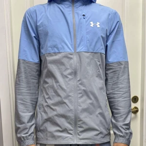 Under Armour blågrå vindjacka - Snygg tvåfärgad vindjacka från Under Armour i ljusblått och grått. Jackan har huva, dragkedja framtill och en diskret logga på bröstet. Perfekt för dig som gillar sportig stil och vill ha något lätt och smidigt till vardags. Modellen är 187