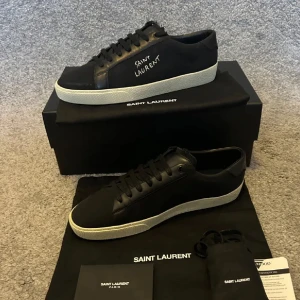 Svarta Saint Laurent sneakers  - Snygga svarta sneakers från Saint Laurent med vit sula och handskriven logga på sidan. Skorna är i skinn med klassisk rund tå och svarta snören. Perfekta för dig som gillar stilrena och exklusiva sneakers med en clean vibe. Orderbekräftelse finns! 