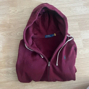Vinröd hoodie från Polo Ralph Lauren - Säljer en vinröd hoodie från Polo Ralph Lauren med dragkedja, huva och vita snören. Klassisk broderad logga på bröstet och två fickor framtill. Perfekt för dig som gillar en chill och stilren look.
