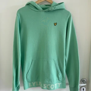 Mintgrön hoodie från Lyle & Scott - Fräsch mintgrön hoodie från Lyle & Scott med klassisk gul logga på bröstet och stor ficka framtill. Tröjan har huva med snörning och ribbade muddar. LYLE & SCOTT står tryckt längs nederkanten. Perfekt för chill dagar eller när du vill sticka ut lite extra.
