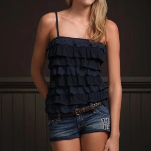 Mörkblå  hollister topp  - Supersöt mörkblå linnetopp med tunna axelband och flera lager volanger som ger en cool och lekfull look. Perfekt för varma dagar när du vill sticka ut lite extra. Snygg att matcha med shorts eller kjol.