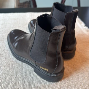 Svarta chelsea boots från Axel Arigato - Stilrena svarta chelsea boots från Axel Arigato i blankt skinn med elastiska paneler på sidorna och diskret logga i guld vid hälen. Klassisk rund tå och platt sula gör dem både snygga och bekväma till alla outfits.