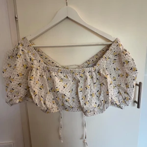 Vit blommig croppad bolerotopp - Supersöt vit topp med små gula blommor och svarta prickar. Toppen har puffiga korta ärmar och snörning i ryggen. Croppad passform med resår i midjan och ärmarna. Perfekt för sommarens alla tillfällen🍋