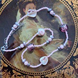 Rosa och lila pärlhalsband med snäckor - Unikt halsband med små rosa glaspärlor, oregelbundna lila och grå pärlor samt skimrande snäckskal och stora lila glaspärlor. Fina kontraster mellan pärlorna och snäckorna ger ett coolt och lekfullt uttryck. Silverfärgat lås.