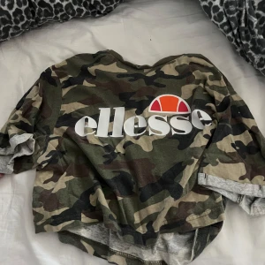 Camouflage t-shirt från Ellesse - Snygg t-shirt från Ellesse med klassiskt camouflagemönster i gröna och bruna toner. Stor logga framtill i vitt och orange. Kortärmad och i mjuk bomull, perfekt för en chill streetwear-look.