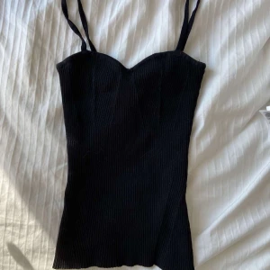 Svart ribbad korsett-topp - Snygg svart korsett-topp med smala axelband och ribbad struktur. Toppen har en figurnära passform och en lätt hjärtformad urringning. Perfekt att styla med jeans eller kjol för en trendig look.