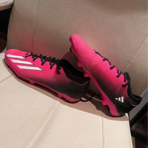 Adidas rosa och svarta fotbollsskor - Snygga fotbollsskor från Adidas i en grymt cool rosa och svart färgkombination med vita ränder på sidan. Skorna har snörning, mönstrad ovandel och dobbar för bästa grepp på planen. Perfekta för dig som vill sticka ut på matchen!