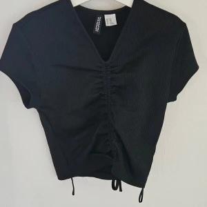 Svart ribbad croppad topp Divided - Svart ribbad croppad topp från Divided med v-ringning och snygg snörning framtill. Kortärmad modell i stretchigt material som sitter tajt och har dragsko-detalj i midjan. Perfekt för att lyfta din outfit med en trendig touch.