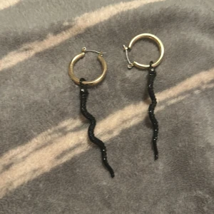 Guldiga hoops med svarta ormhängen - Coola örhängen med guldiga hoops och svarta, glittriga ormhängen. Ormarna är smala och vågiga, vilket ger en edgy vibe. Perfekta för dig som vill sticka ut med ett unikt smycke. Kan ta av ormar 