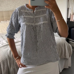 Randig blåvit blus med knappar - Snygg blus med blå och vita ränder, knappar framtill och lätt rynkade ärmslut. Luftig modell med trekvartsärm och rund halsringning. Perfekt för en avslappnad och stilren look.