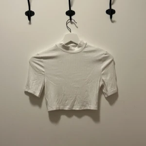 Vit croppad ribbad topp - En stilren vit croppad topp med ribbad struktur och korta ärmar. Toppen har en högre halsringning och är perfekt för att matcha med höga jeans eller kjol. Enkel och clean design som passar till många olika looks.