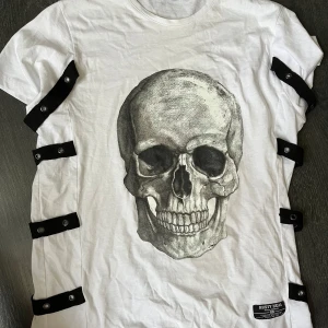 Vit t-shirt med svarta band Rusty Neal - Säljer en vit t-shirt från Rusty Neal med unika svarta band och tryckknappar längs sidorna. T-shirten har rund hals, korta ärmar och ett tryck framtill. Materialet är mjuk bomull och passformen är normal. Perfekt för dig som vill sticka ut lite extra.