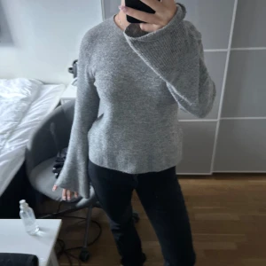 Grå stickad tröja med vida ärmar - Mysig grå stickad tröja med rund hals och långa, vida ärmar. Tröjan har en enkel och stilren design med ribbade muddar vid ärmslut och nederkant. Perfekt för lager-på-lager och chill dagar.