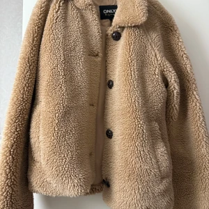Beige teddyjacka från ONLY - Supermjuk och fluffig teddyjacka från ONLY i beige färg. Jackan har klassisk krage, stora knappar framtill och är helfodrad. Perfekt för dig som vill ha en mysig och trendig look under kyliga dagar.