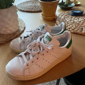 Adidas Stan Smith vita sneakers - Klassiska Adidas Stan Smith sneakers i vitt skinn med gröna detaljer på hälen och tungan. Lågt skaft, perforerade ränder på sidorna och platt sula. Perfekta för en clean och tidlös stil. Ikonisk logga på hälen och tungan.