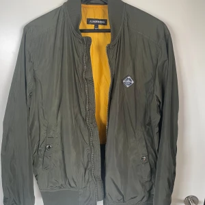 Olivgrön bomberjacka från J.Lindeberg - Snygg olivgrön bomberjacka från J.Lindeberg med gul insida och klassisk ribbad krage. Jackan har dragkedja framtill, två fickor med knapp och en patch på bröstet. Perfekt för dig som vill ha en stilren och trendig look.