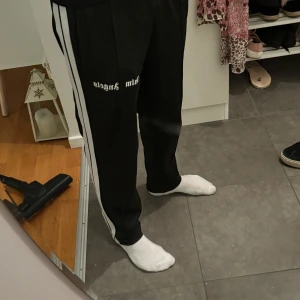 Palm Angels svarta mjukisbyxor - Svarta mjukisbyxor från Palm Angels med vita sidoränder och logga tryckt på låret. Byxorna har en relaxed passform och elastisk midja. Perfekta för en chill och sportig look.