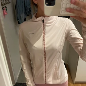 Ljusrosa träningsjacka från SOC - Säljer en ljusrosa träningsjacka från SOC med hel dragkedja framtill och diskret logga på bröstet. Jackan har lång ärm, smal passform och är tillverkad i ett lätt syntetmaterial som andas. Perfekt för löprundan eller powerwalken.